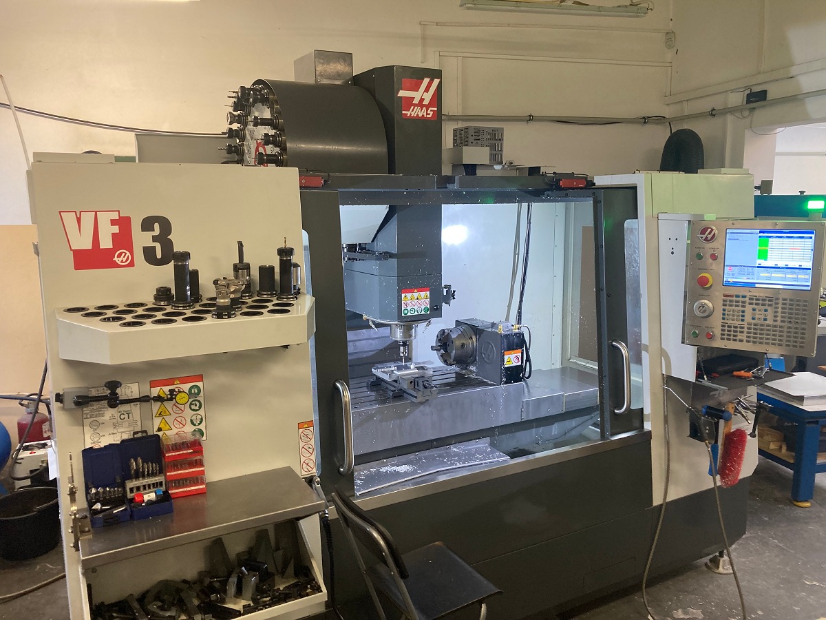 Wnętrze warsztatu z maszyną CNC Haas VF-3, widoczne narzędzia skrawające, panel sterowania z ekranem, obrabiany element w uchwycie, oświetlenie robocze i elementy wyposażenia stanowiska.