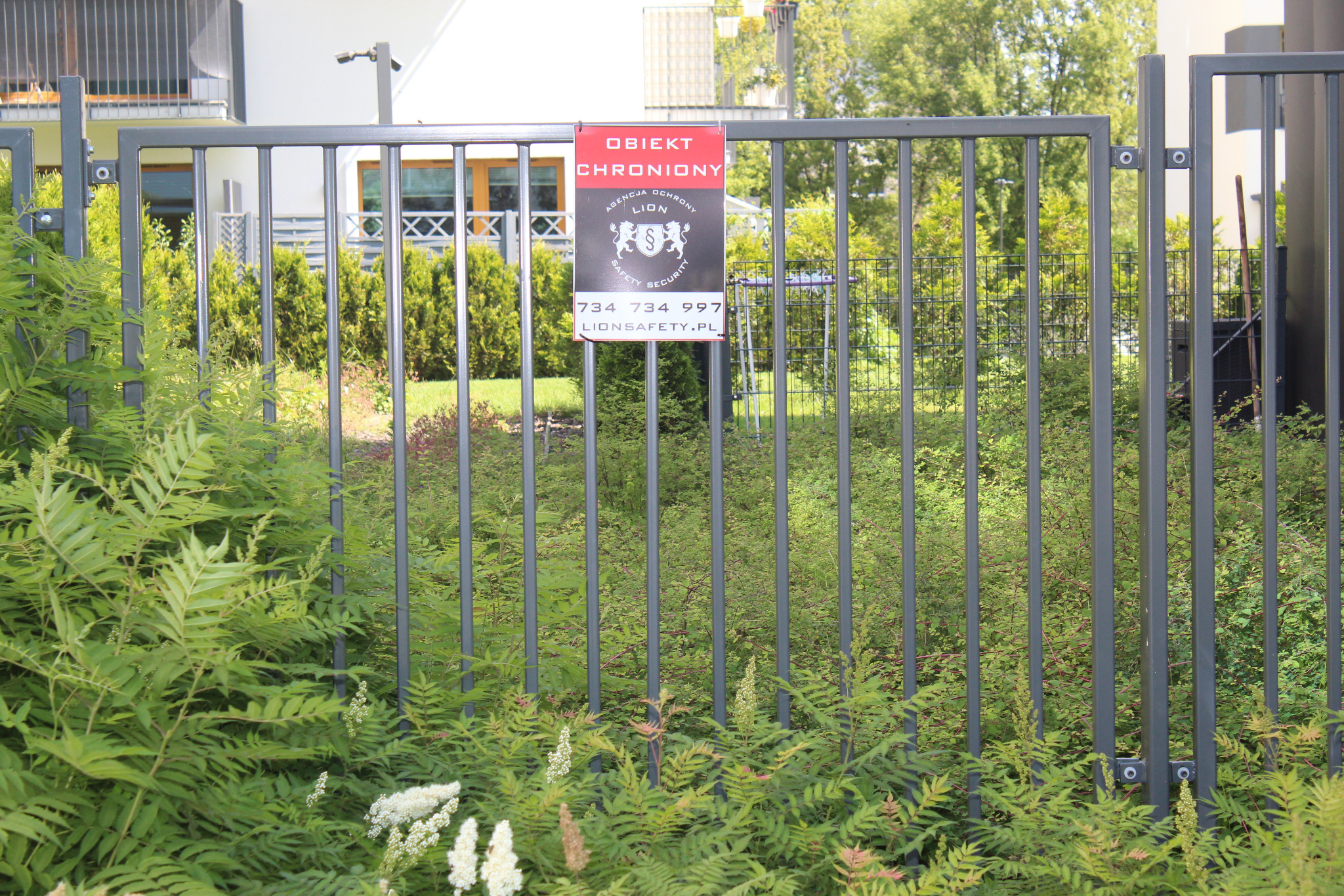 Tabliczka informacyjna 'Obiekt chroniony' agencji ochrony Lion, przymocowana do metalowego ogrodzenia posesji, z widoczną zielenią w tle.