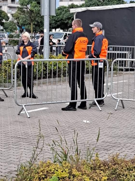 Trzech pracowników ochrony w czarnych spodniach i pomarańczowo-czarnych kurtkach z napisem 'LION' i 'Służby Porządkowe' stoi za metalowymi barierkami na zewnątrz.