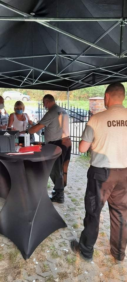 Ochroniarze w szarych koszulkach z napisem 'OCHRONA' obsługują klientów przy czarnym stoliku pod zadaszeniem, kobieta w maseczce przegląda dokumenty.