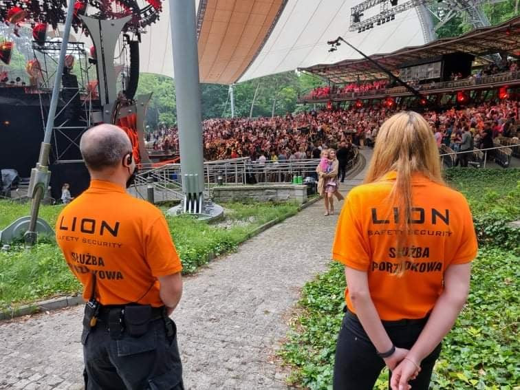 Dwóch pracowników służby porządkowej w pomarańczowych koszulkach z napisem 'LION SAFETY SECURITY' stoi tyłem do kamery, obserwując duży tłum ludzi w amfiteatrze podczas wydarzenia.