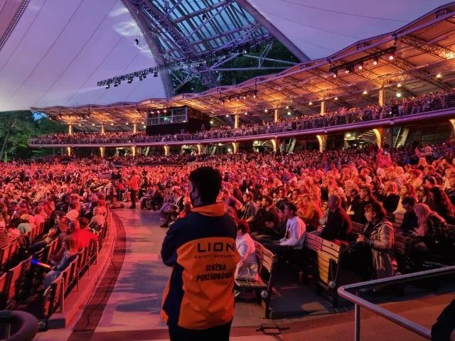 Ochroniarz w pomarańczowo-granatowej kurtce z napisem 'Służba Porządkowa' stoi tyłem do kamery na tle amfiteatru wypełnionego publicznością oświetloną czerwonym światłem.