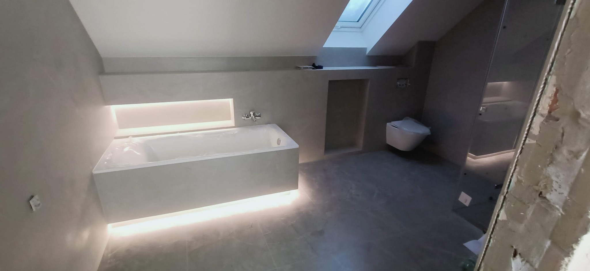 Nowoczesna łazienka w Bargłówce w trakcie remontu, z podświetlaną wanną, wiszącą toaletą i oknem dachowym, widoczne niedokończone prace przy kabinie prysznicowej.
