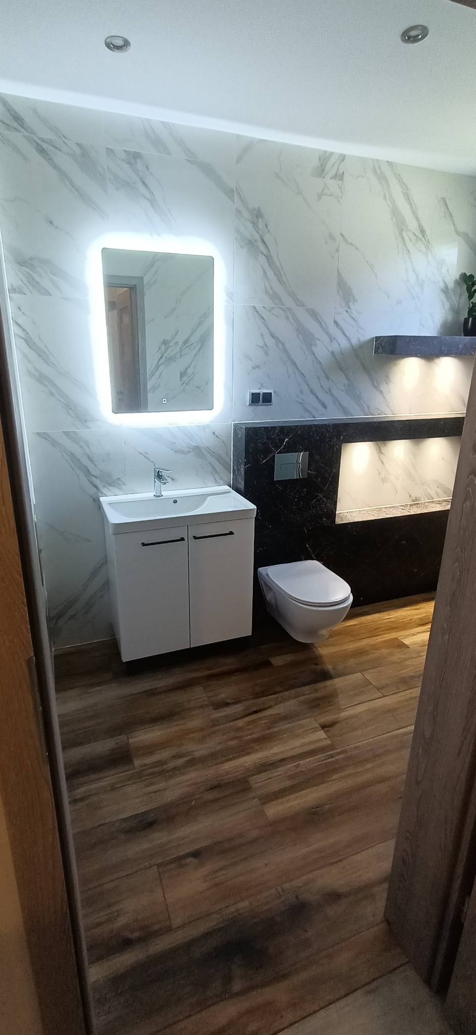 Nowoczesna łazienka z podświetlanym lustrem, marmurowymi płytkami na ścianach, drewnopodobną podłogą i podwieszaną toaletą.