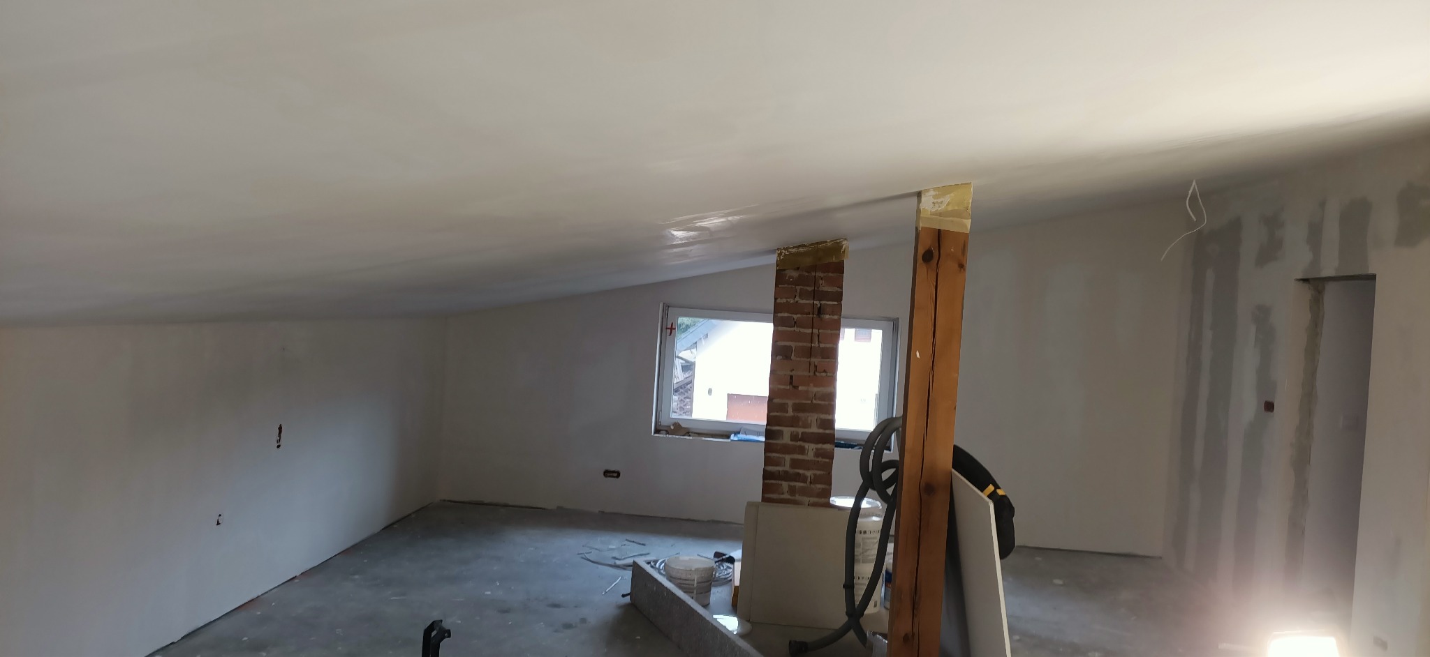 Wnętrze w trakcie remontu: gładź na skośnym suficie, ceglana kolumna z taśmą zabezpieczającą, drewniana belka, ściany z widocznymi śladami szpachlowania, okno z widokiem na zewnątrz.