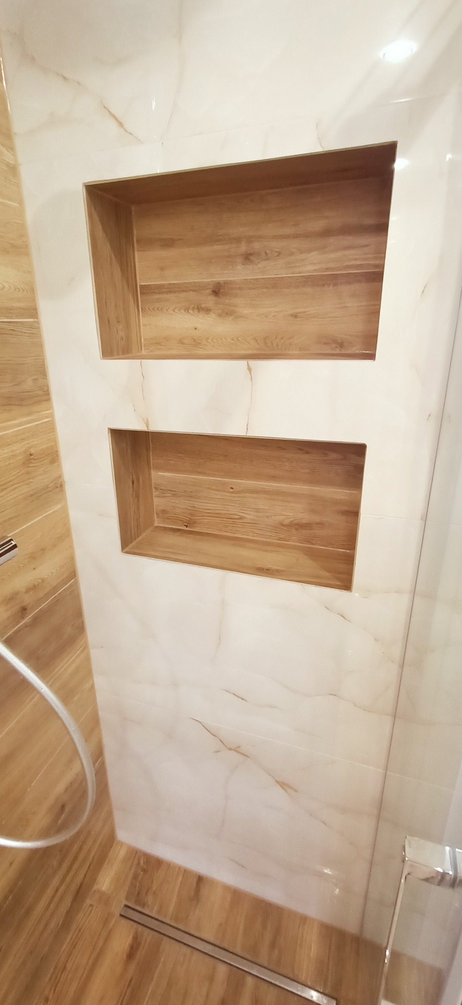 Dwa podświetlone, prostokątne wnęki w ścianie wyłożonej marmurkowymi płytkami, z drewnopodobnym wykończeniem wnętrza wnęk, widoczny fragment kabiny prysznicowej i słuchawki.