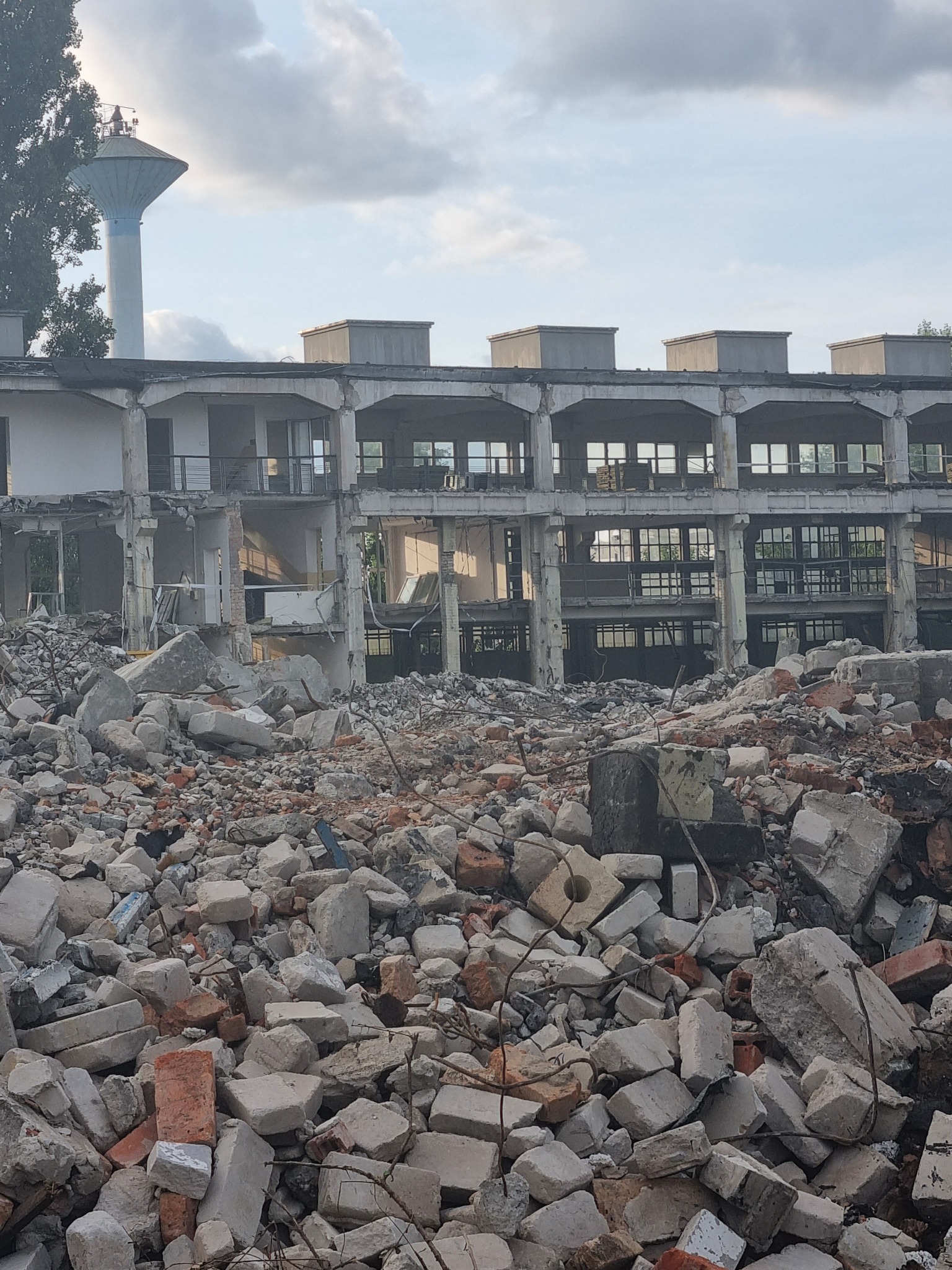Zdjęcie przedstawia gruzowisko po rozbiórce budynku, z widocznymi pozostałościami ścian i kolumn, na tle błękitnego nieba z chmurami i wieżą ciśnień w oddali.