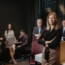 Zespół prawny w biurze: kobieta z blond włosami na pierwszym planie, skrzyżowane ręce, za nią czterech współpracowników w eleganckich strojach, nowoczesne biuro z żaluzjami w tle.