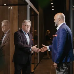 Dwóch mężczyzn w garniturach w trakcie uścisku dłoni w nowoczesnym biurze z przeszklonymi ścianami i drewnianymi elementami dekoracyjnymi, w tle widoczna kobieta.