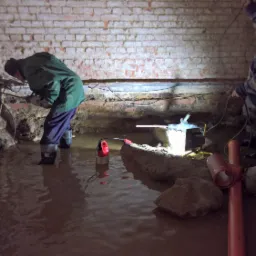 Dwóch pracowników w gumowych butach, w zalanym, ceglanym pomieszczeniu, prawdopodobnie piwnicy, jeden wierci w ścianie, drugi trzyma kabel, w wodzie widoczna pompa i rury PCV.