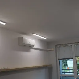 Wnętrze biura lub mieszkania po remoncie, widoczne liniowe lampy LED na suficie, klimatyzator na ścianie, okno z żaluzjami i półki wzdłuż ścian.