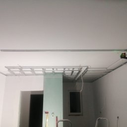 Usługi remontowo - budowlane - Montaż stelażu sufitu podwieszanego z użyciem poziomicy laserowej, widoczna płyta gipsowo-kartonowa i fragment okna.