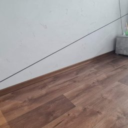 Usługi remontowo - budowlane - Nowoczesne wnętrze z panelami podłogowymi w odcieniach brązu, białą ścianą z dekoracyjną fakturą i czarnymi liniami, oraz szarą szafką RTV z minimalistycznym wazonem.