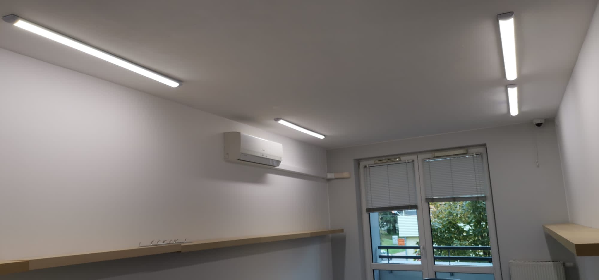 Wnętrze biura lub mieszkania po remoncie, widoczne liniowe lampy LED na suficie, klimatyzator na ścianie, okno z żaluzjami i półki wzdłuż ścian.