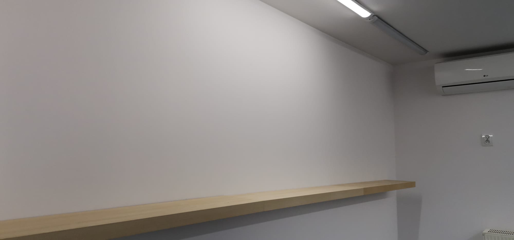 Gładka, pomalowana na biało ściana z zamontowaną, surową, drewnianą półką, klimatyzacją LG i gniazdkiem elektrycznym, oświetlona podłużną lampą sufitową.