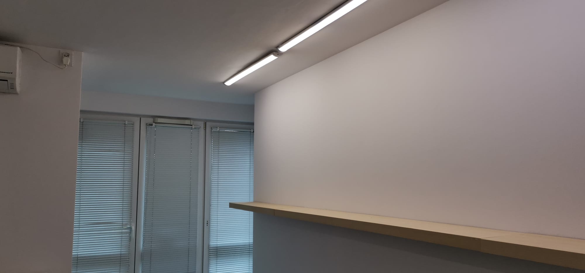 Wnętrze pokoju po remoncie z gładką, pomalowaną na biało ścianą, drewnianą półką, oknami z białymi żaluzjami i liniowym oświetleniem LED na suficie.