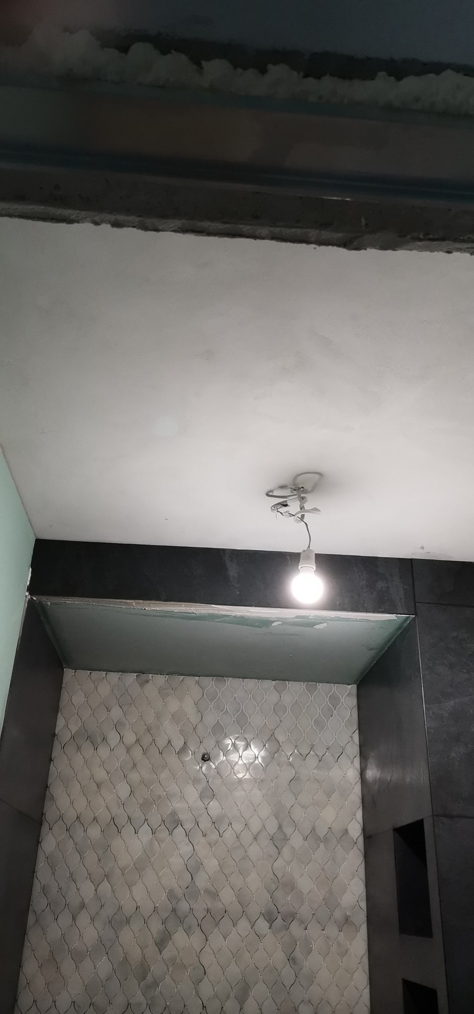 Wnętrze łazienki w trakcie remontu: widoczna mozaika na ścianie, ciemne płytki, surowy sufit z prowizorycznym oświetleniem żarówką.