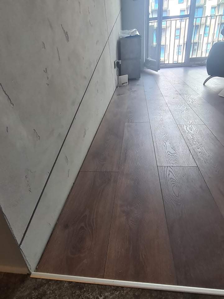 Ukończona podłoga z paneli drewnopodobnych, widok z niskiego kąta, listwa przypodłogowa w kolorze aluminium, ściana z szarą dekoracyjną strukturą, duże okno balkonowe w tle.