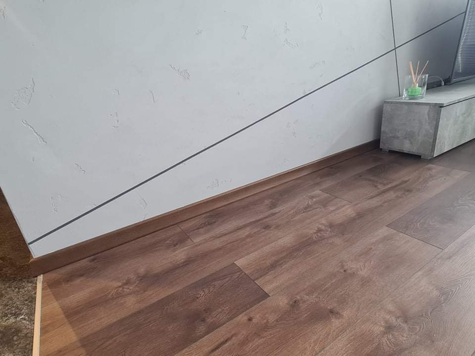 Nowoczesne wnętrze z panelami podłogowymi w odcieniach brązu, białą ścianą z dekoracyjną fakturą i czarnymi liniami, oraz szarą szafką RTV z minimalistycznym wazonem.