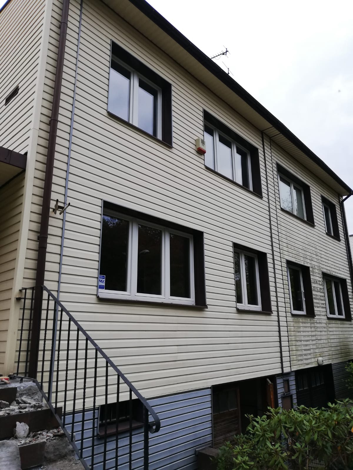 Dwupiętrowy budynek z sidingiem w odcieniach beżu i brązu, z widocznymi oknami z białymi ramami i czarnymi obramowaniami, oraz fragmentem schodów z czarną balustradą. Część elewacji jest zabrudzona...