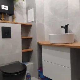 Nowoczesna łazienka z szarą marmurową okładziną, drewnianym blatem pod umywalką nablatową i czarną armaturą, widoczny wiszący sedes i bojler z rośliną.