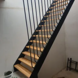 Nowoczesne schody drewniane z czarną metalową balustradą w surowym wnętrzu, widoczny kubeł z farbą Śnieżka i drabinka w tle.