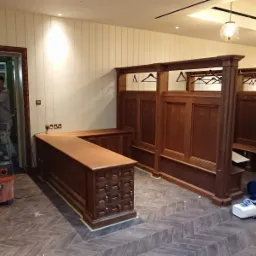 Wykończone drewniane wnętrze szatni z blatem i wieszakami, widoczny pracownik sprzątający po montażu, podłoga w jodełkę.