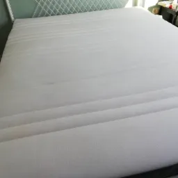 Świeżo wyprany, biały materac z delikatnymi śladami po czyszczeniu widocznymi pod odpowiednim kątem światła, leżący na ł&oacute;żku z metalowym zagł&oacute;wkiem.