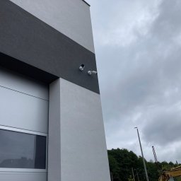 AURUM IT sp. z o.o. - Dwie kamery monitoringu zamontowane na szarej ścianie budynku z czarnym pasem, widok z dołu na pochmurne niebo i fragment koparki w tle.