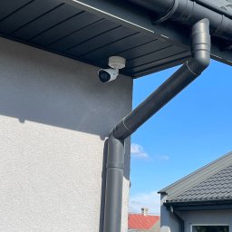 Esafer.pl - Kompleksowe Systemy Zabezpieczeń - Biała kamera IP zamontowana pod ciemnym okapem domu z szarą elewacją. Widoczny fragment rynny i błękitne niebo w tle. Zabezpieczenie domu.