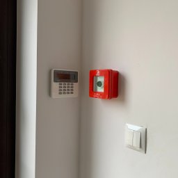 AURUM IT sp. z o.o. - Panel alarmowy Satel, czerwona obudowa przeciwpożarowa z młotkiem i włącznik światła na białej ścianie, obok drzwi z kluczami. Ujęcie z bliska.
