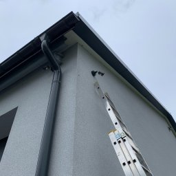 Esafer.pl - Kompleksowe Systemy Zabezpieczeń - Kamera monitoringu zamontowana na elewacji domu, widoczna drabina oparta o ścianę. Ujęcie z dołu, pochmurne niebo w tle. Szara elewacja i orynnowanie.