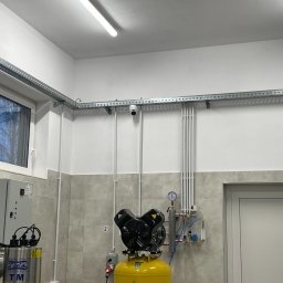 Esafer.pl - Kompleksowe Systemy Zabezpieczeń - Kamera monitoringu zamontowana na ścianie w pomieszczeniu przemysłowym, widoczne koryta kablowe, instalacje rurowe i kompresor. Szaro-białe płytki na ścianach.