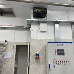 AURUM IT sp. z o.o. - Instalacja przemysłowa: szafa sterownicza, kable w korytkach, drabina oparta o ścianę z planami. Widoczne elementy instalacji technologicznej i elektrycznej.