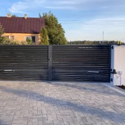 Nowoczesna, automatyczna brama wjazdowa z poziomych, czarnych profili, z widocznymi siłownikami i centralą sterującą, na tle domu z brązowym dachem i kostki brukowej w odcieniach szarości.