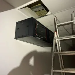 Czarna szafa serwerowa zamontowana na ścianie w pomieszczeniu biurowym, z widocznym otwartym sufitem podwieszanym i aluminiową drabiną opartą o ścianę.