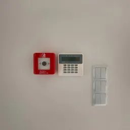 Czerwony przycisk alarmowy pożarowy Elektromet, biała klawiatura sterująca systemem alarmowym Safetel Perfecta oraz potrójny włącznik światła zamontowane na białej ścianie w pobliżu okna.