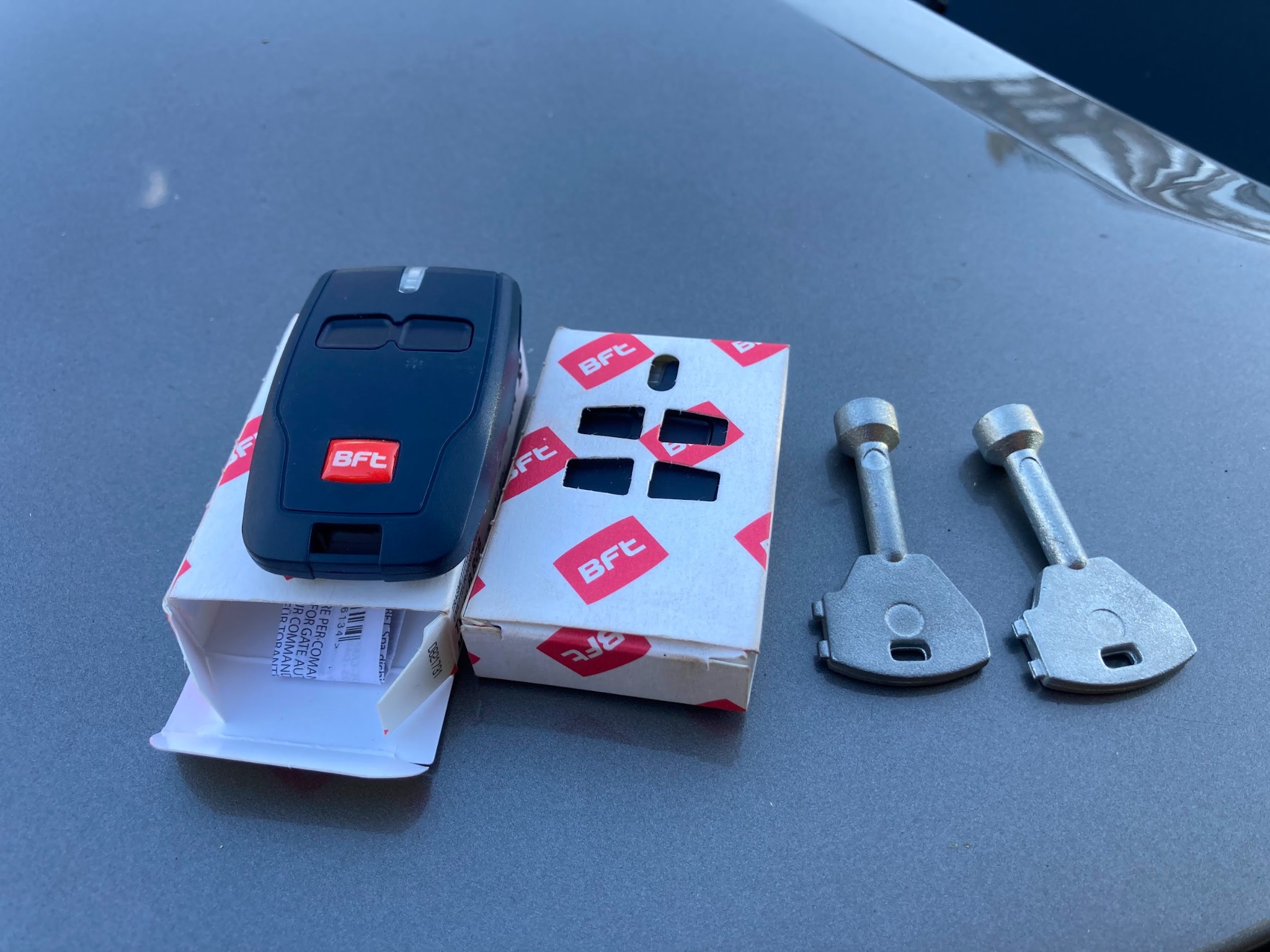 Zestaw startowy BFT: pilot do bramy automatycznej na pudełku obok dwóch kluczy do siłownika, leżący na szarym tle.
