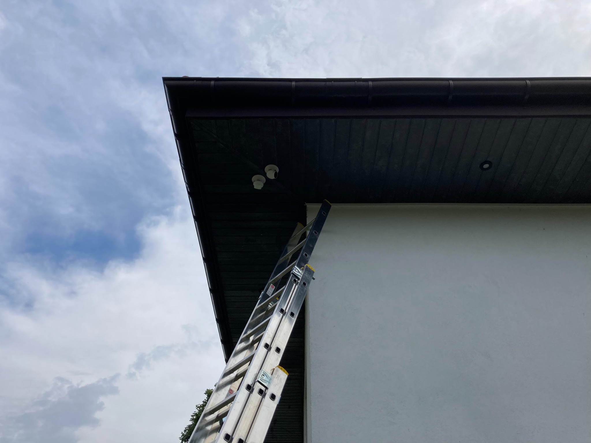 Aluminiowa drabina oparta o białą ścianę domu, pod okapem z zamontowanymi dwoma białymi kamerami monitoringu.