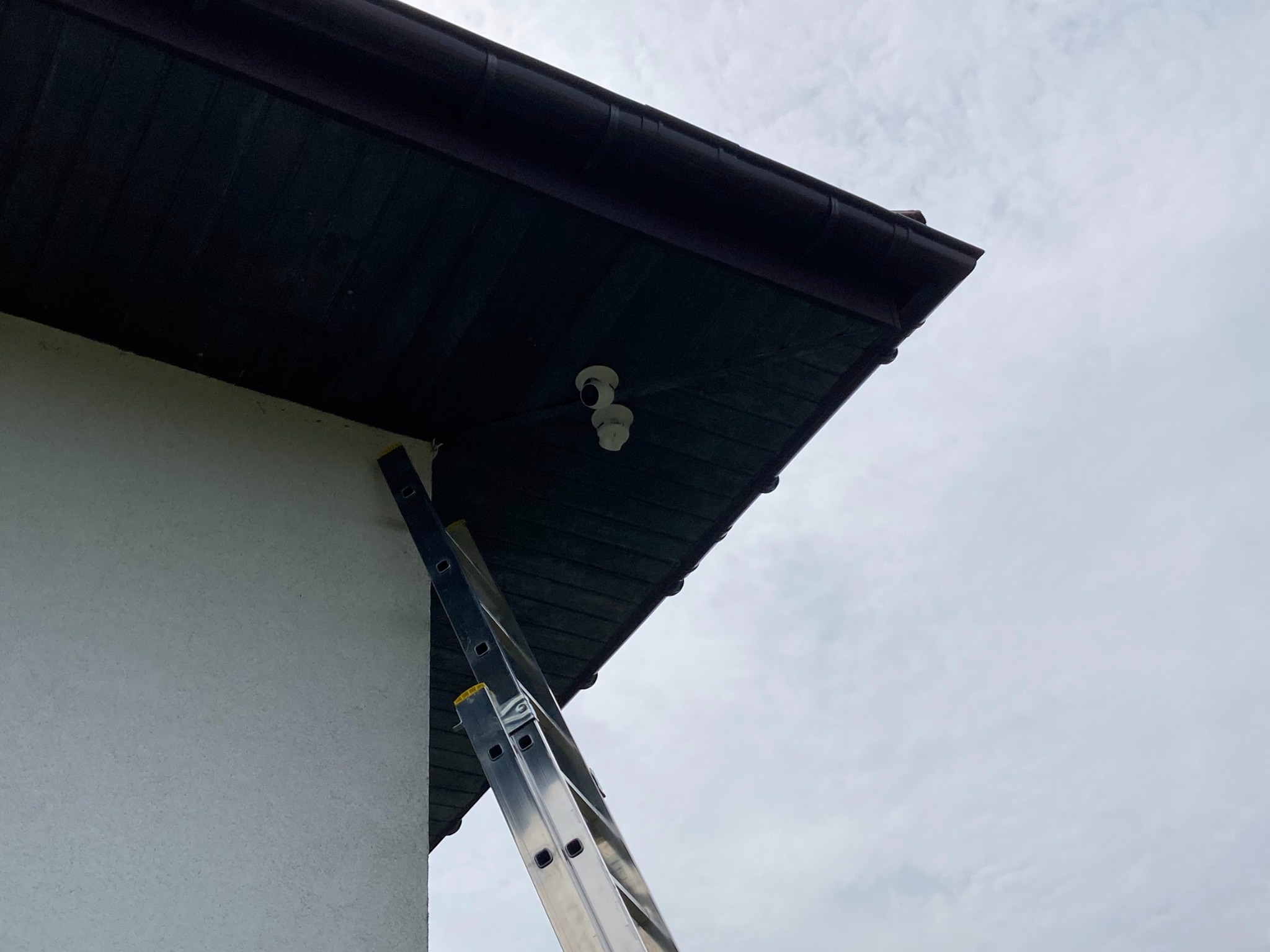 Dwie białe kamery monitoringu zamontowane pod okapem budynku, widoczne z dołu z częściowo rozłożoną aluminiową drabiną opartą o ścianę.