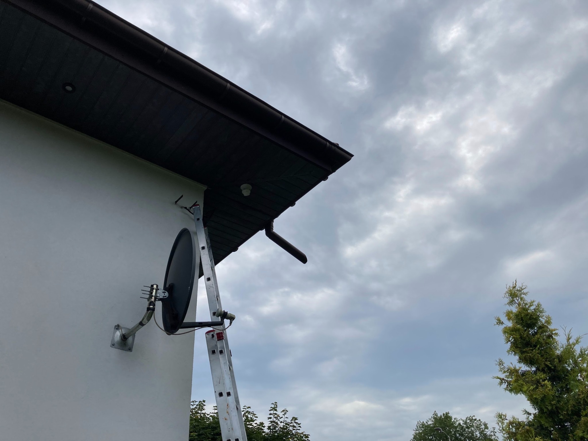 Antena satelitarna zamontowana na ścianie domu, widoczna drabina oparta o ścianę, pochmurne niebo w tle.