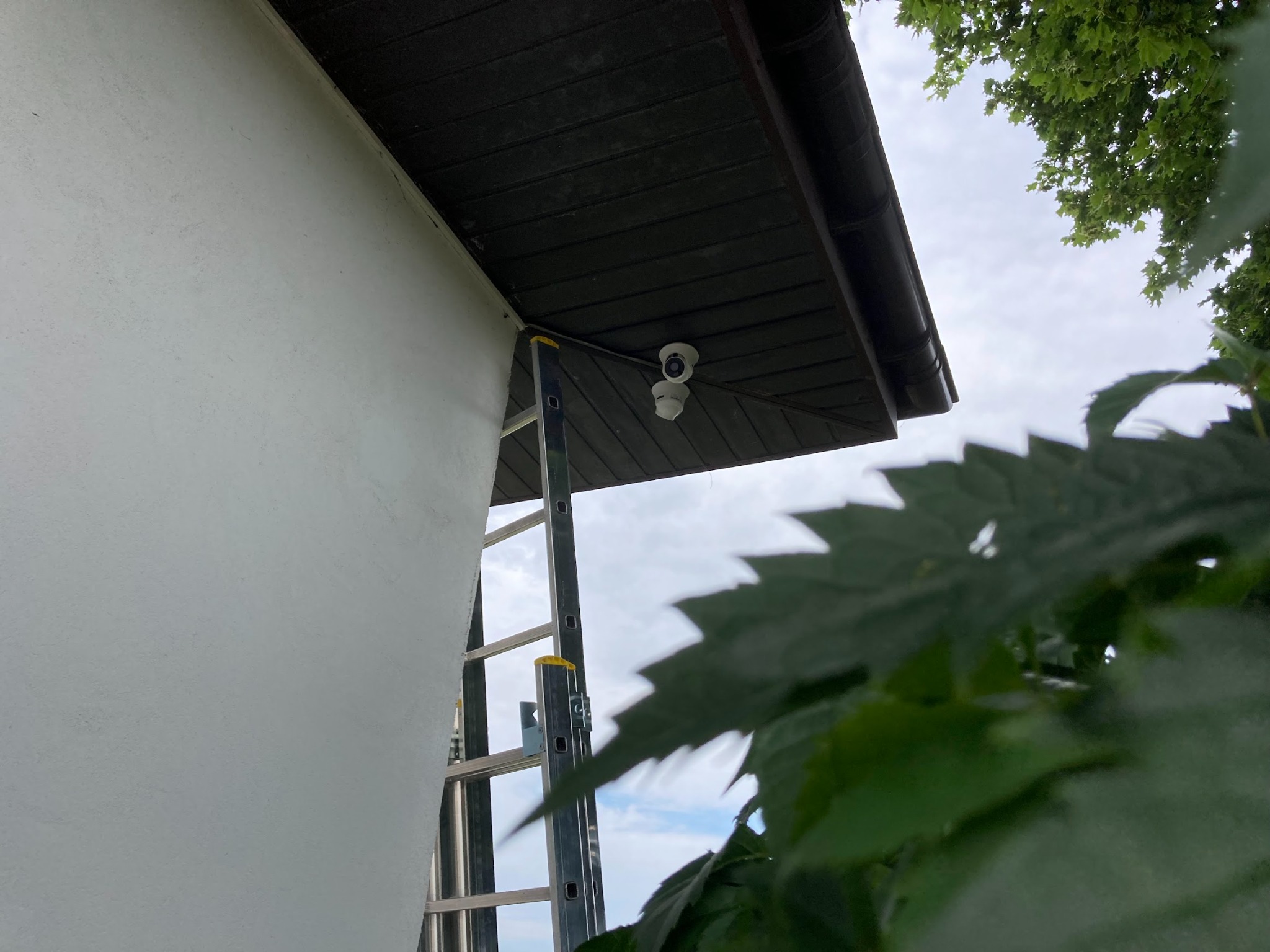 Dwie kamery monitoringu zamontowane pod okapem domu, widoczne z dołu z użyciem drabiny, częściowo zasłonięte przez liście.