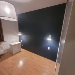 Pokój po remoncie z ciemnogranatową ścianą, drewnianą podłogą, białymi lampkami ściennymi i białym regałem, widok z perspektywy narożnika.