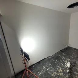 Świeżo pomalowane, jasnoszare ściany w pokoju, oświetlone lampą roboczą na tr&oacute;jnogu; podłoga zabezpieczona folią malarską, widoczny otw&oacute;r w suficie.