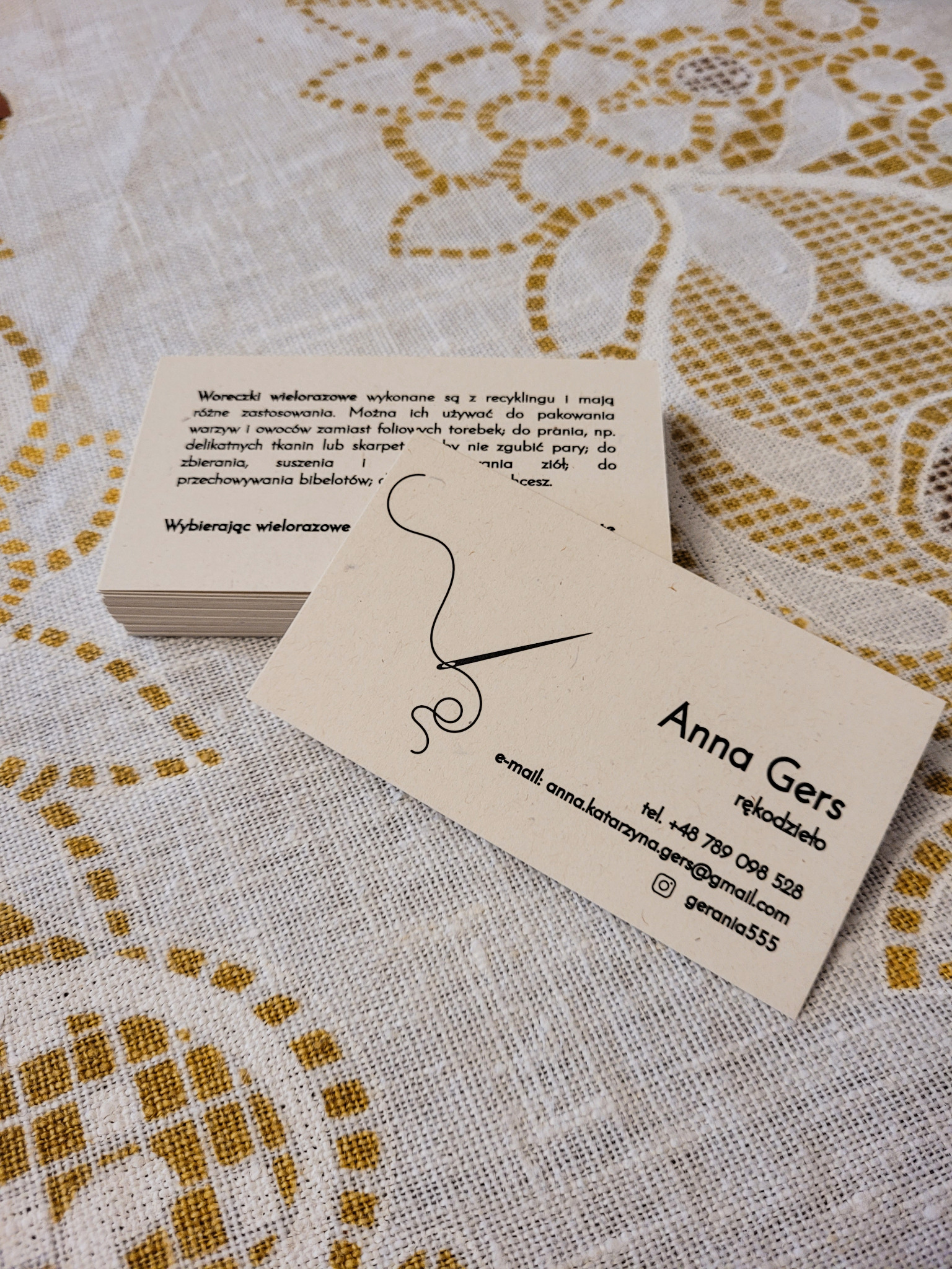 Stos wizytówek i jedna wizytówka z logotypem igły i nitki na tle lnianej serwety. Wizytówki 'Anna Gers rękodzieło' z danymi kontaktowymi.