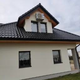 Boczny widok na dom z zainstalowaną jednostką klimatyzacji Haier na poddaszu, czarna dachówka i okna z żaluzjami.