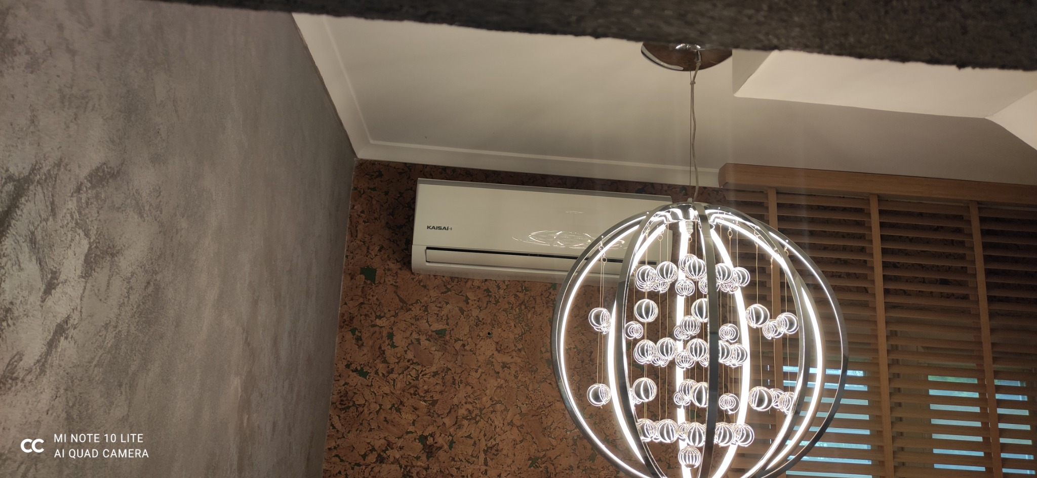 Biały klimatyzator Kaisai zamontowany na korkowej ścianie w pomieszczeniu z dekoracyjnym oświetleniem kulistym i drewnianymi żaluzjami.