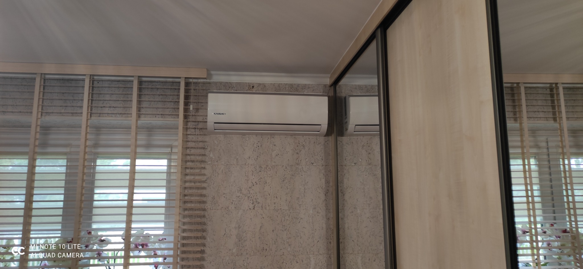 Biały klimatyzator marki Kaisai zamontowany na ścianie w pokoju z żaluzjami i przesuwną szafą z lustrem.