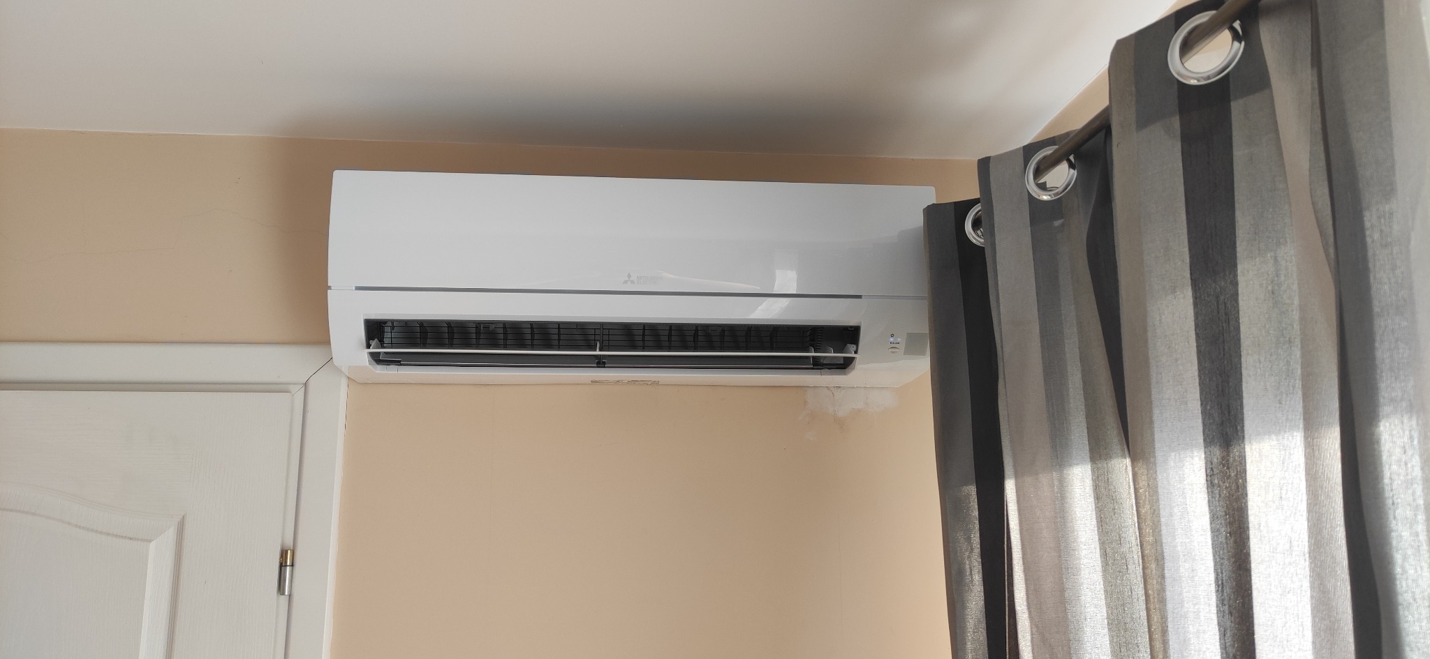 Biała klimatyzacja Mitsubishi zamontowana na beżowej ścianie w pokoju, widoczne szare zasłony i białe drzwi.