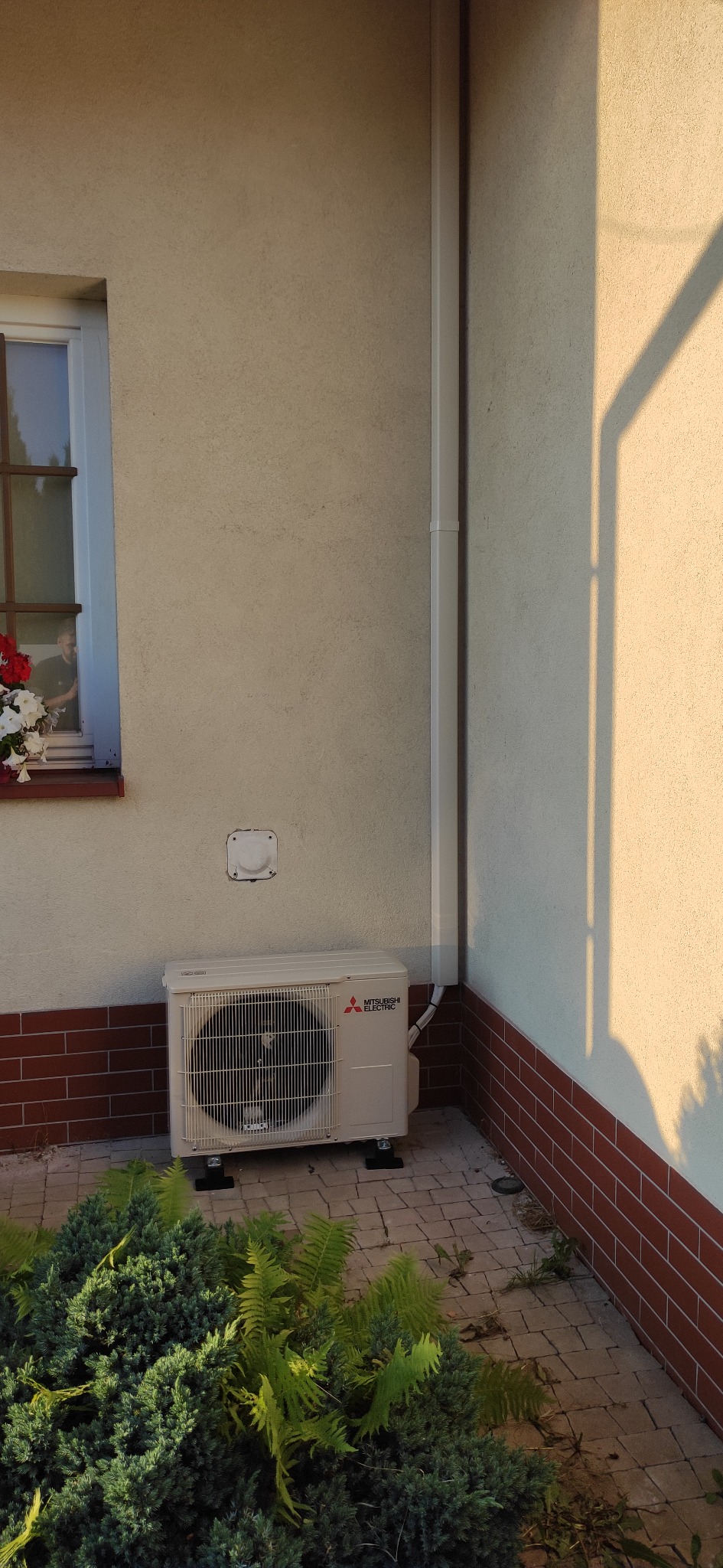 Zewnętrzna jednostka klimatyzacji Mitsubishi Electric zamontowana przy ścianie domu z ceglaną podmurówką, widoczna zieleń dekoracyjna i cień padający na elewację.